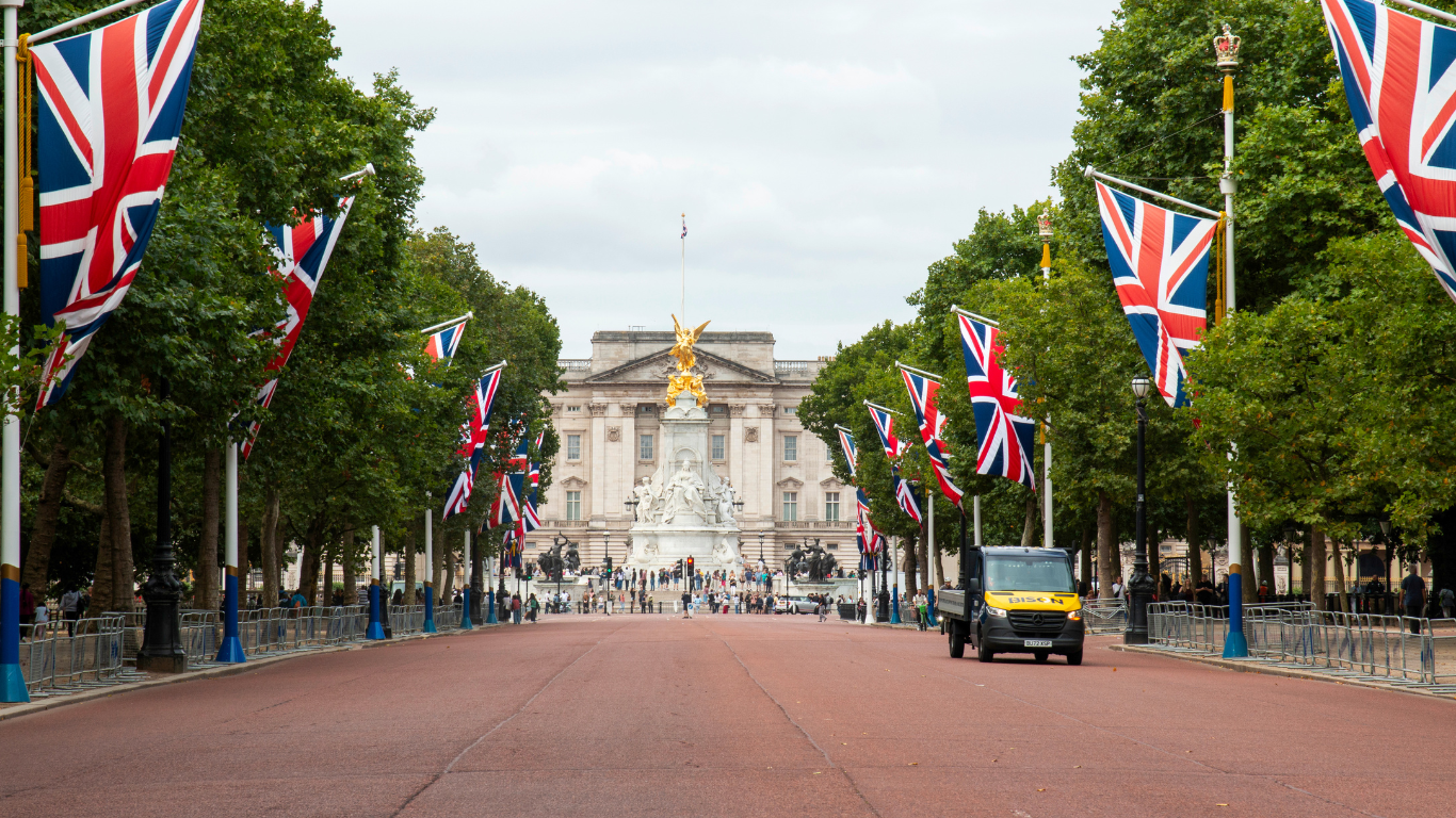 Allée bordée de drapeaux britanniques, arbres verts et monument central devant un bâtiment historique à Londres.