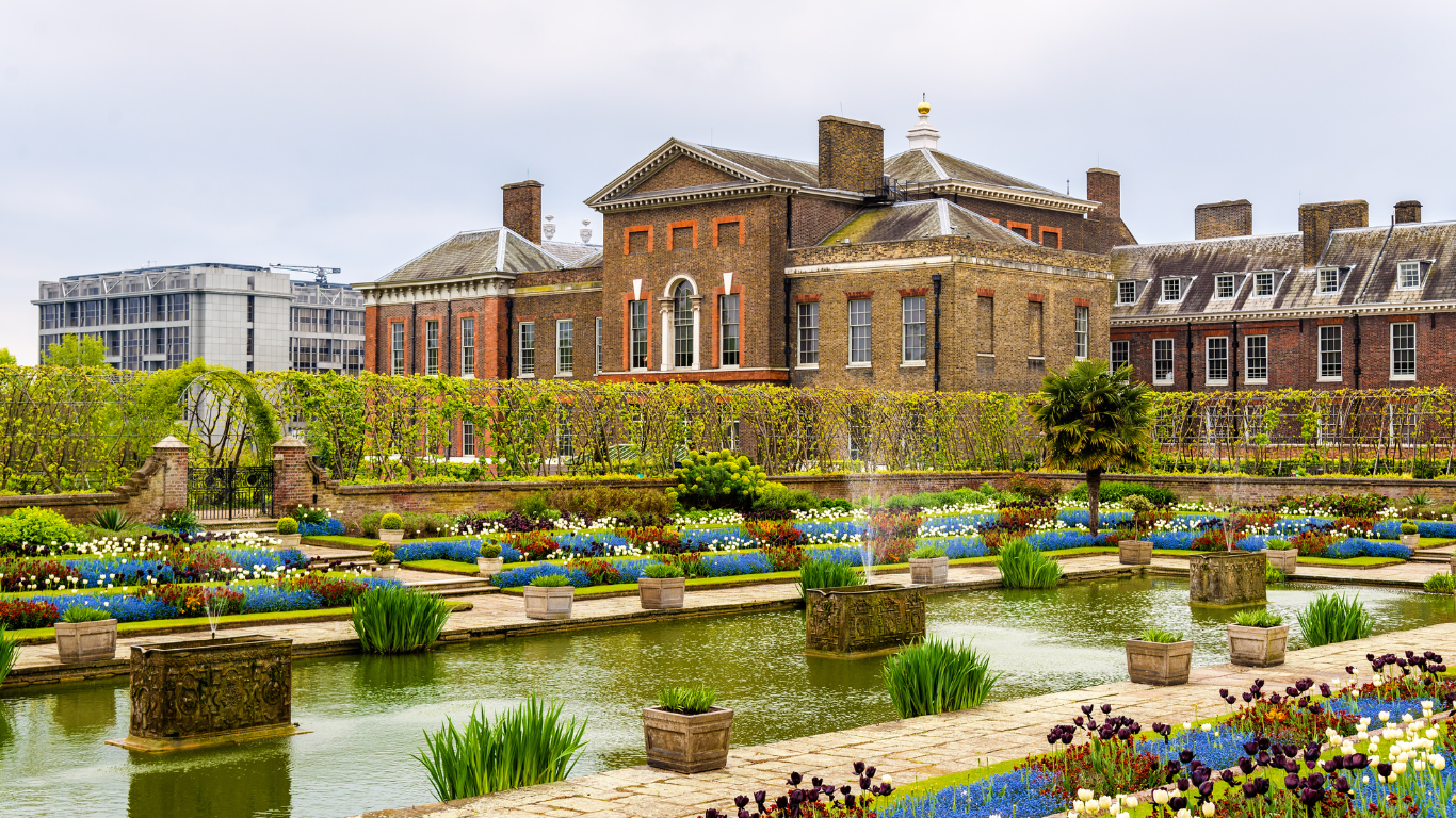 Jardin fleuri avec bassin d’eau, pots en pierre, et bâtiments en briques rouges sous ciel gris.
