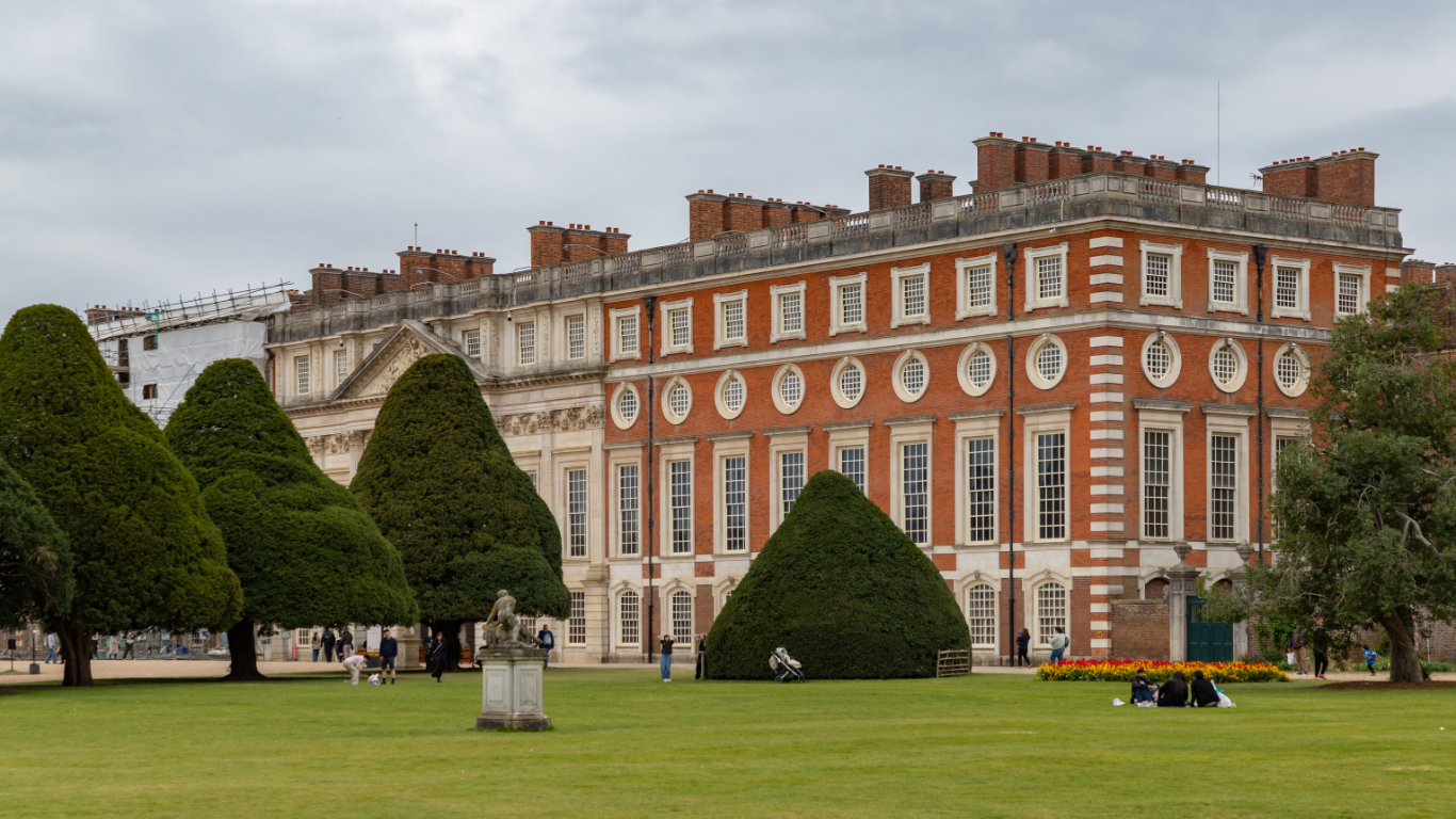 Façade de bâtiment historique en briques et pierre, fenêtres ovales, pelouse verte avec arbres taillés et visiteurs.