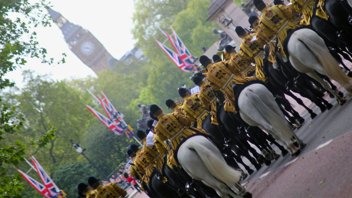 Rangée de chevaux noirs sellés, cavaliers en uniforme jaune, drapeaux britanniques et arbres en arrière-plan.