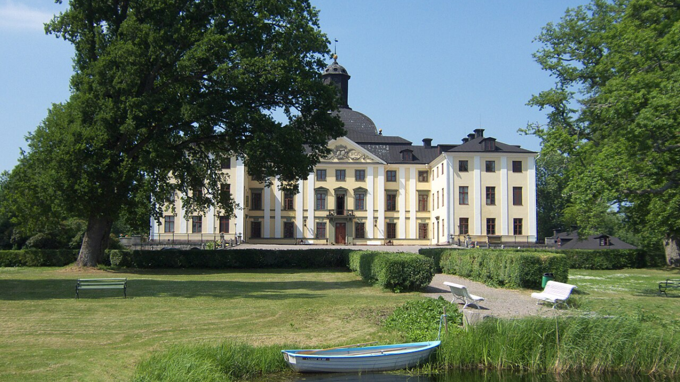 Bâtiment historique blanc avec clocher, entouré d’arbres, pelouse verte et bancs dans un parc.