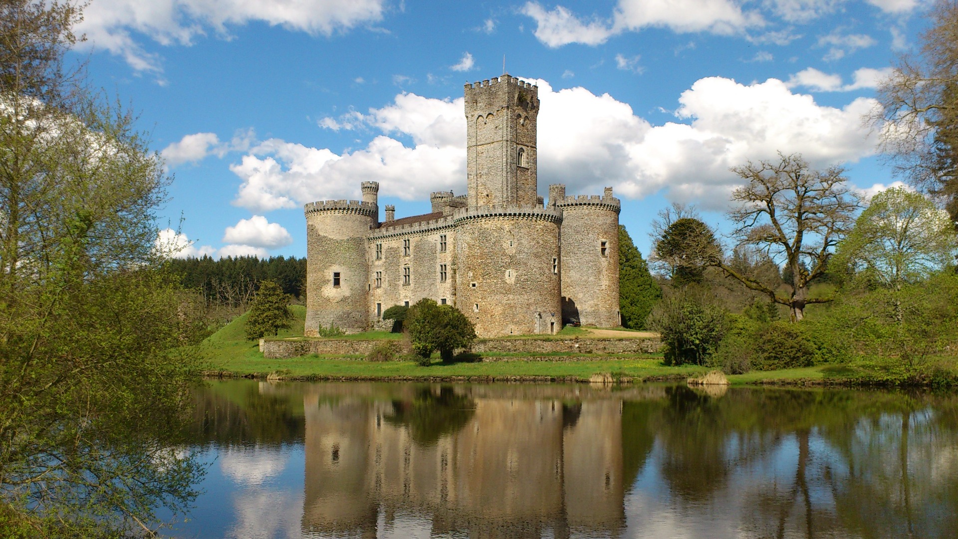 Château médiéval en pierre avec tours, reflet dans un lac, arbres verts et ciel bleu nuageux.