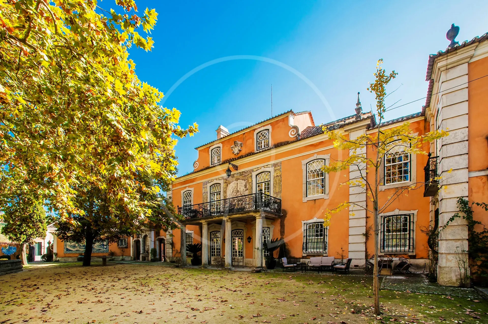 Façade orange et blanche d’un bâtiment historique, fenêtres à barreaux, arbres jaunes et ciel bleu.