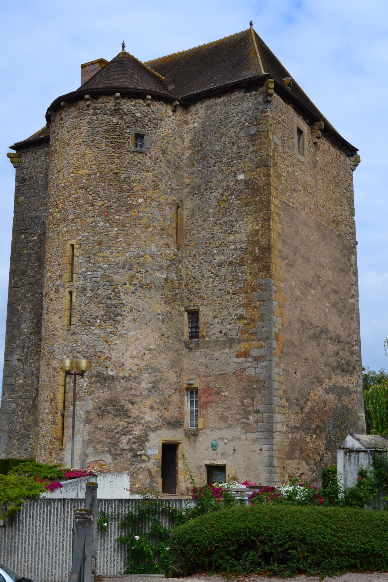 Façade en pierre d’un ancien bâtiment, toit en ardoise, fenêtres étroites, fleurs et jardin au premier plan.