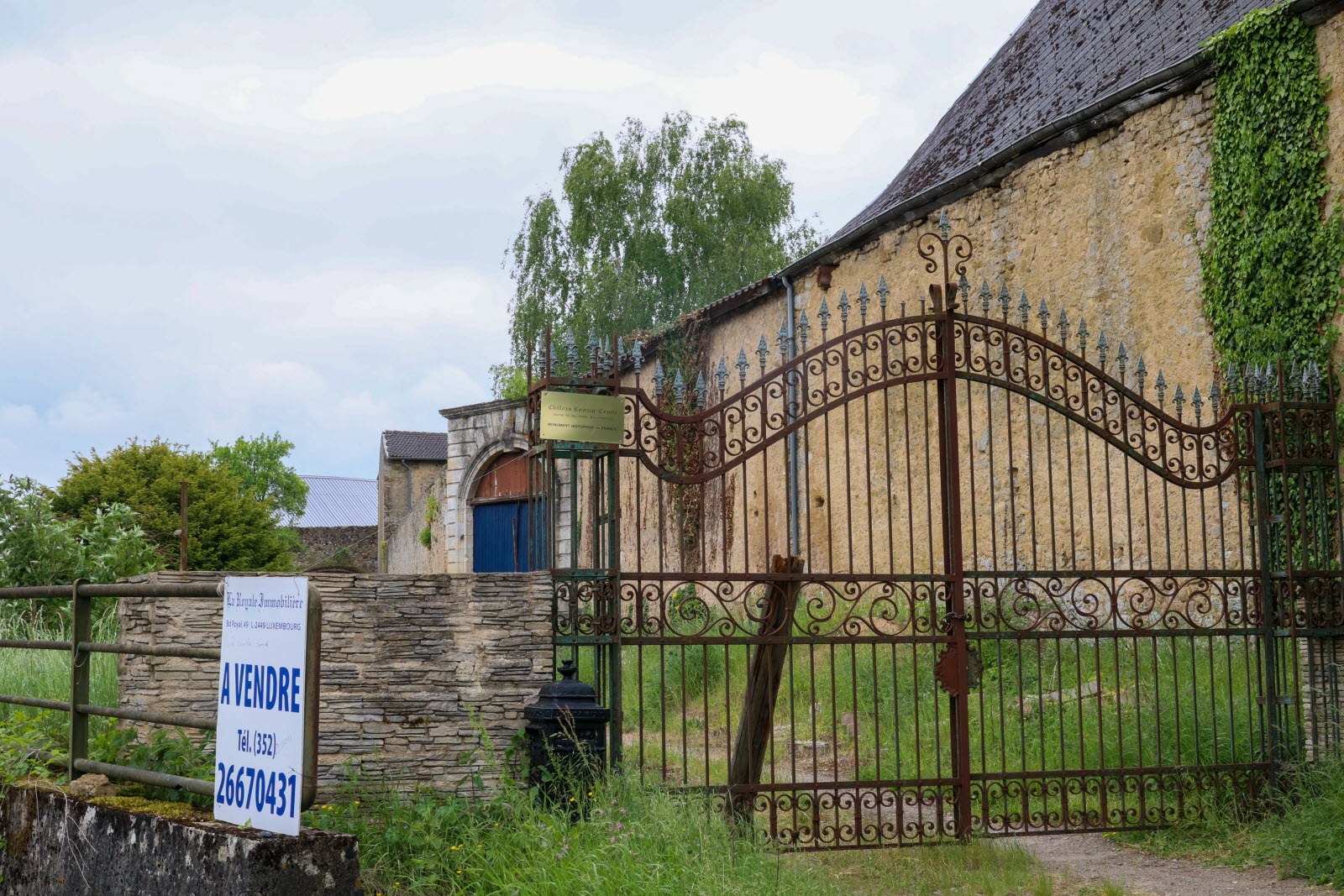 Portail en fer forgé brun devant une grande bâtisse en pierre, jardin vert et ciel nuageux.