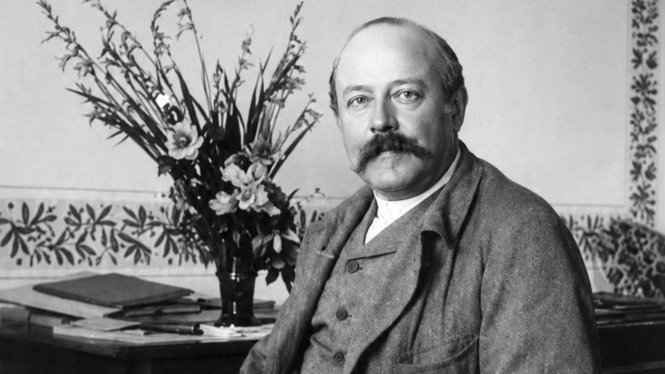 Portrait en noir et blanc d’un homme moustachu assis, près d’un bouquet de fleurs et d’un bureau.