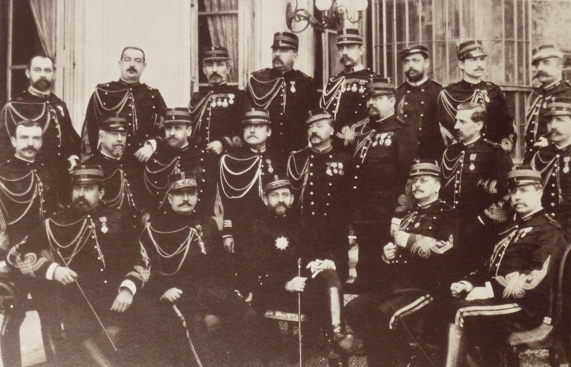Groupe de soldats en uniforme sombre, chapeaux à plumes, posant devant un bâtiment, photo ancienne en noir et blanc.