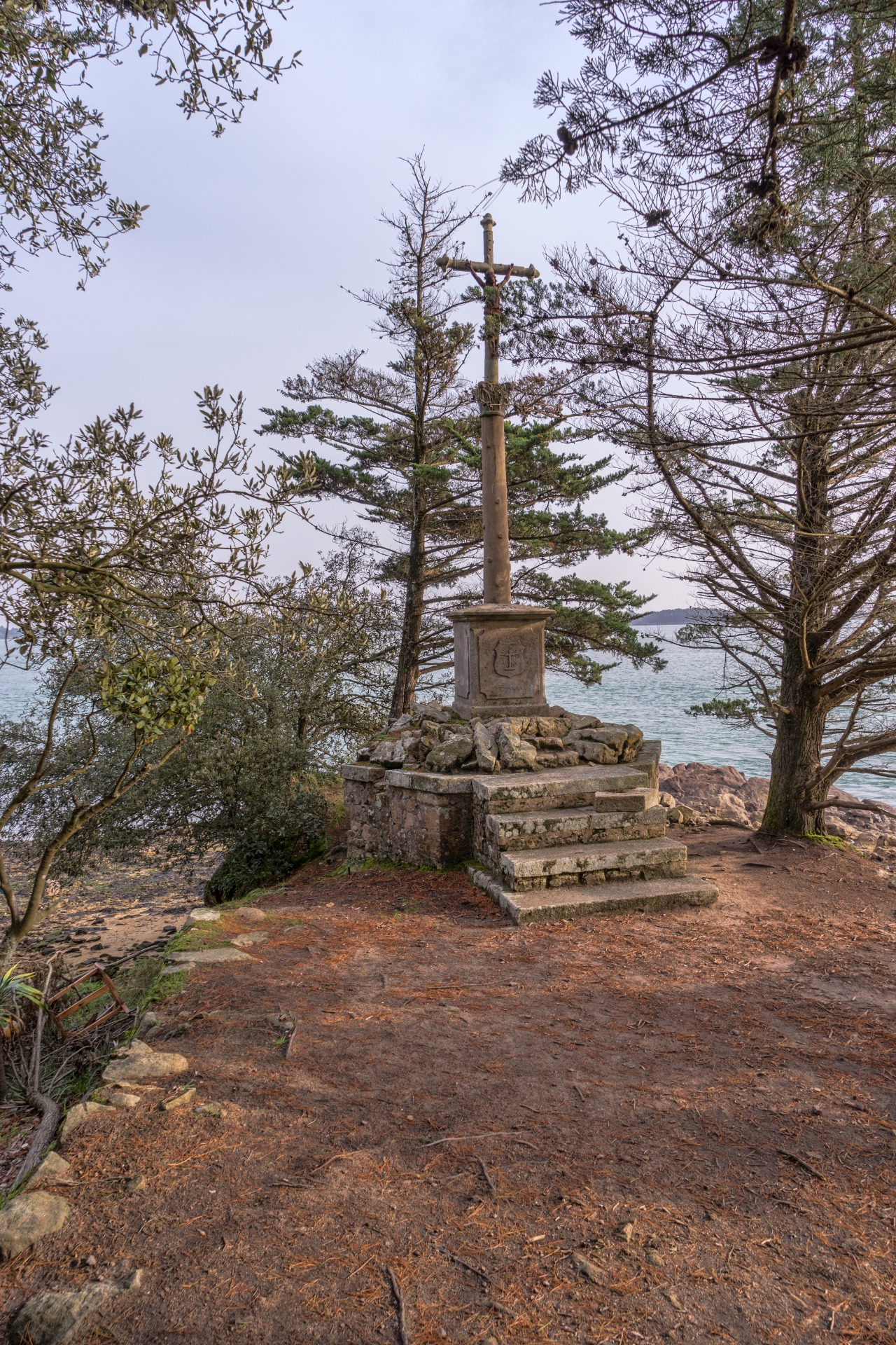 Croix en pierre sur un socle, entourée d’arbres, au bord de la mer sous un ciel gris.