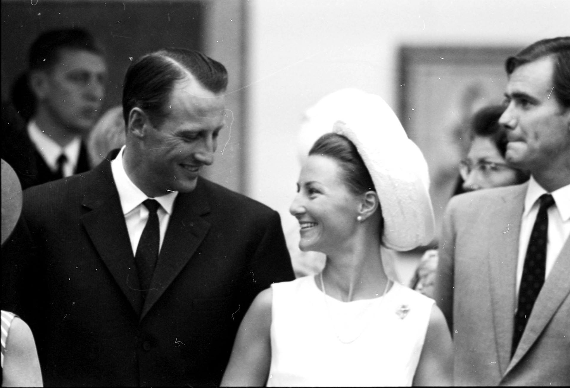 Homme en costume souriant, femme en robe blanche et chapeau, entourés d’autres personnes, photo ancienne.