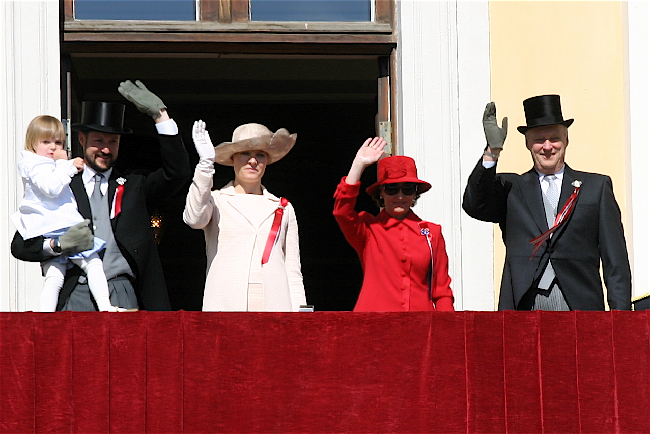 Quatre personnes saluent depuis un balcon, dont un enfant, une femme en chapeau et deux gardes en uniforme rouge.