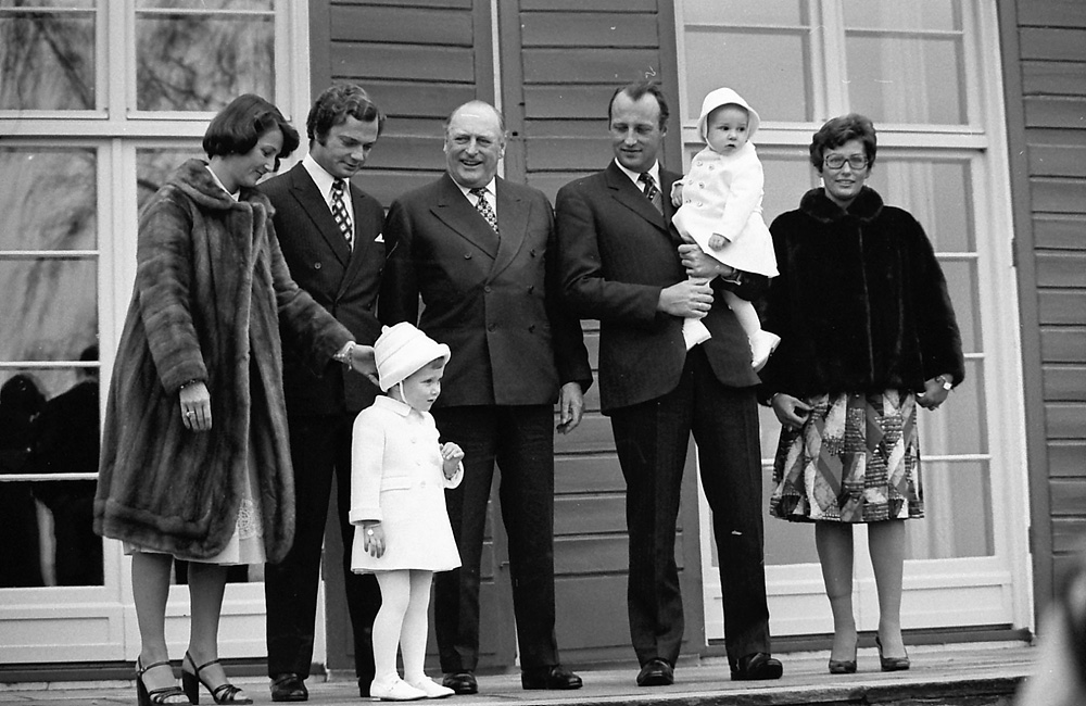 Groupe de personnes devant une maison, un homme tient un bébé, plusieurs adultes et enfants sourient.