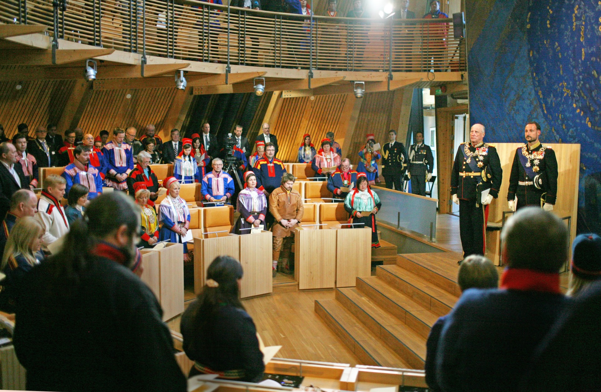 Salle de cérémonie en bois, foule de dignitaires en costumes traditionnels, deux officiers en uniforme décoré, public au premier plan.